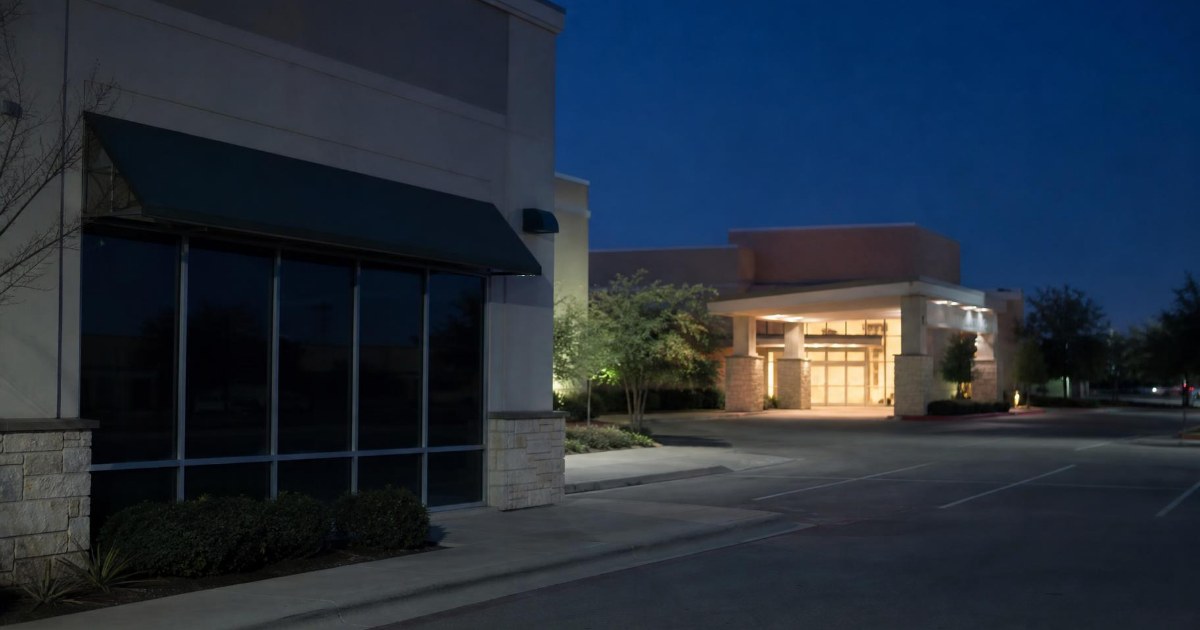 A lit emergency room entrance glowing at night across from a dark, closed clinic storefront.